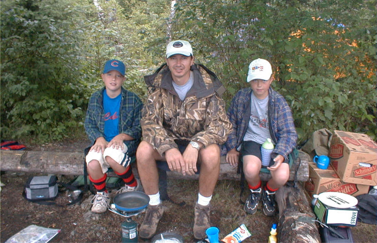 Max Winders with his two sons, Jack and Tommy, sitting on a log at their campsite in the Boundary Waters, surrounded by camping gear and enjoying the wilderness.