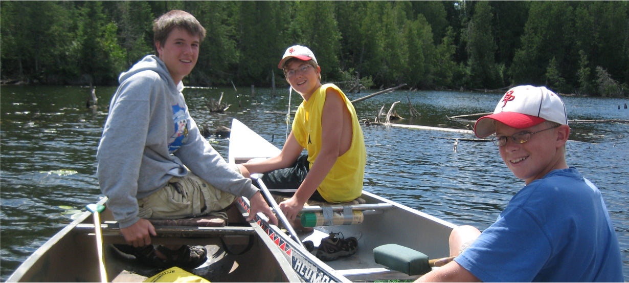 Max Winders' three sons smiling while sitting in a canoe on a serene lake in the Boundary Waters, ready for an adventure.