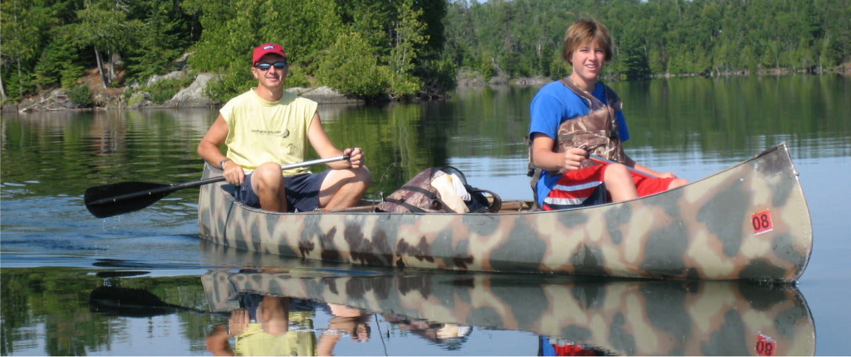 Max Winders and his son, Jeremy, paddling a camouflage-patterned canoe on a calm lake in the Boundary Waters, enjoying a peaceful day on the water.