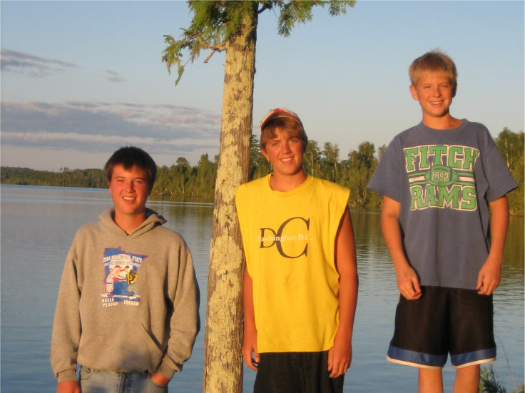 Max Winders' three sons, Jeremy, Tommy, and Jack, standing together by a tree on the shore of a lake in the Boundary Waters, smiling in the golden light of sunset.